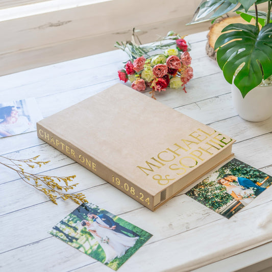 A luxury velvet photo album with gold text on the cover, displayed on a table alongside some photographs and a bouquet of flowers.