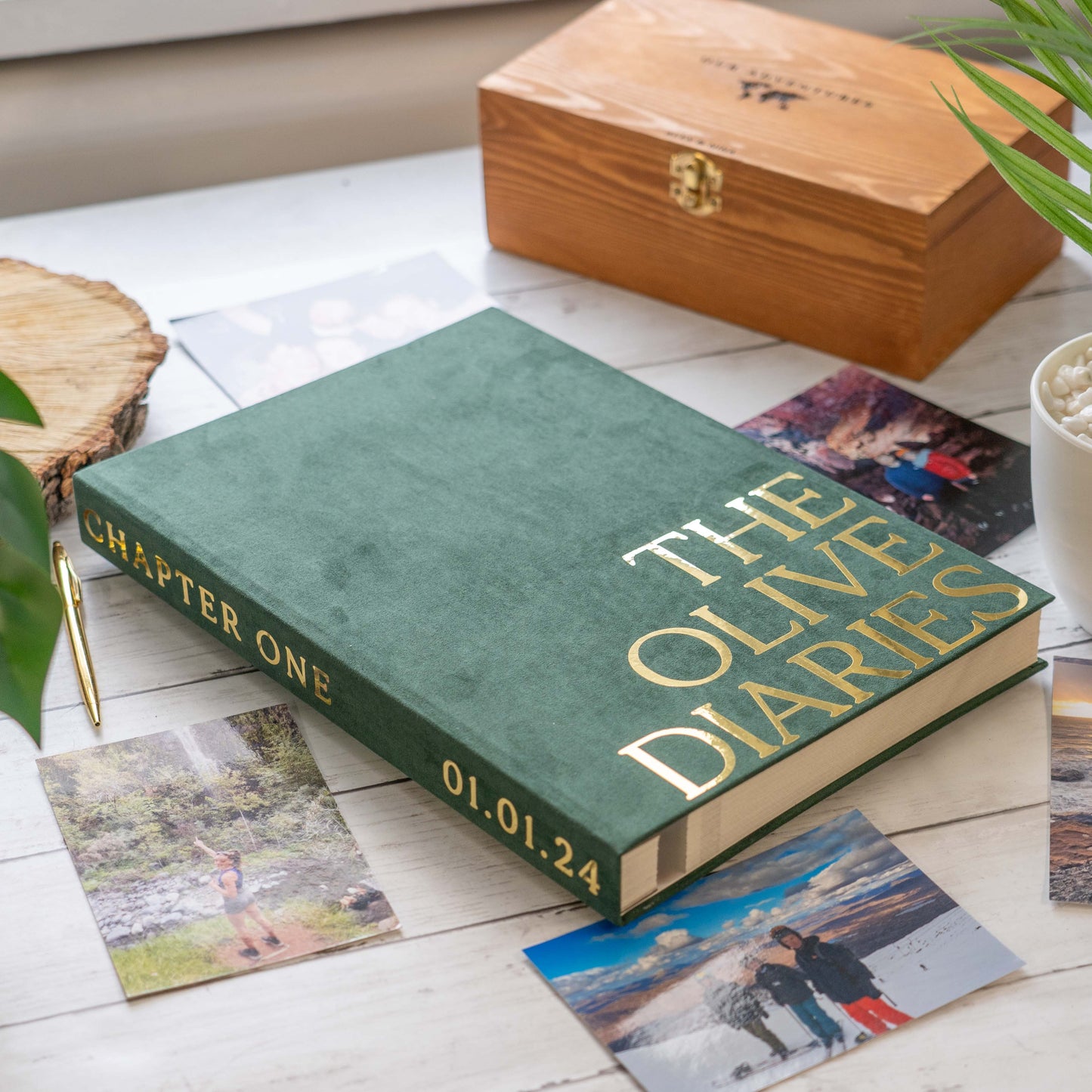 Green book titled 'The Olive Diaries' with gold text on a wooden surface with photos and a wooden box.