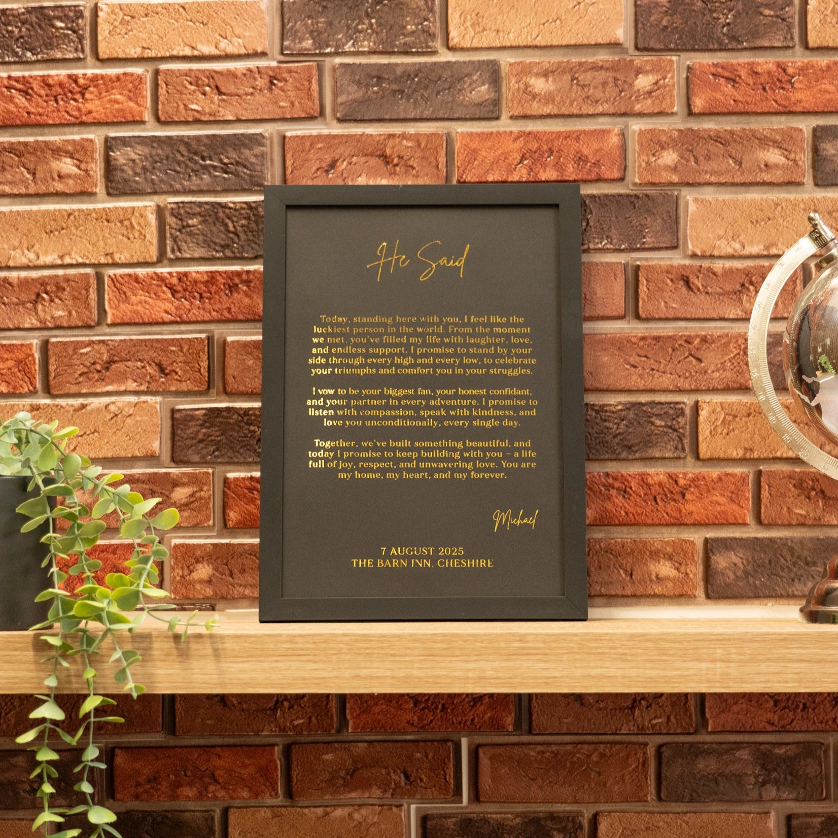 Framed text on a wooden shelf against a brick wall with a plant and decorative object.