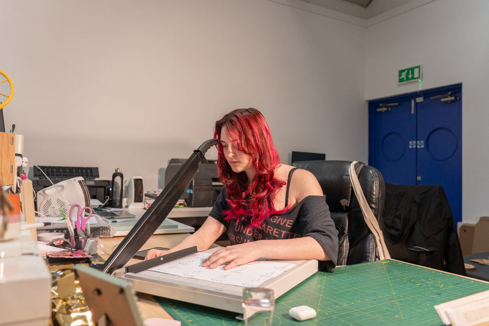 Person with red hair working at a desk with a large paper cutter in an office setting.