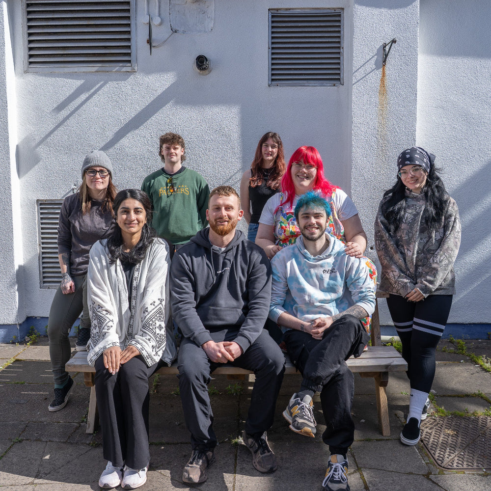 A group of eight young adults posing together outdoors on a bench in front of a white stucco building. Five people are seated on the bench and three stand behind them.
