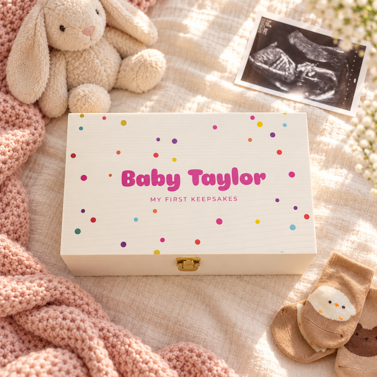 A personalized baby keepsake box with a gold clasp, decorated with colorful confetti dots and the text 'Baby Taylor' and 'MY FIRST KEEPSAKES'. The box is styled in a cozy flat-lay setting with a pink bunny, a baby bootie, and an ultrasound photo on a textured beige blanket.
