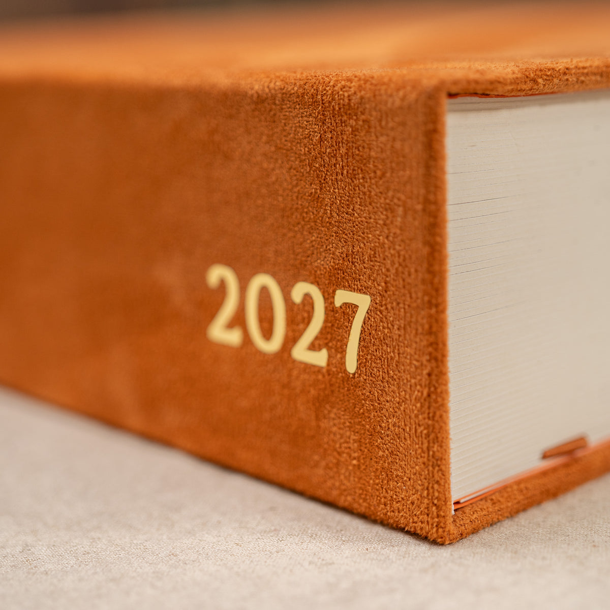 Close-up of a hardcover 2027 journal with a textured orange-brown cover and gold foil year numbering, resting on a neutral surface.
