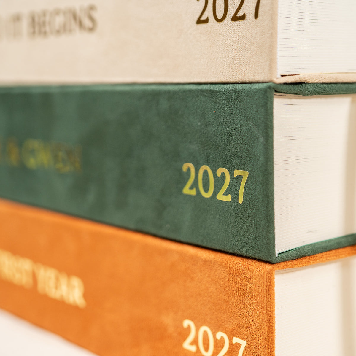 Close-up of stacked hardcover books or journals with textured spines in beige, dark green, and orange, each marked with the year 2027.