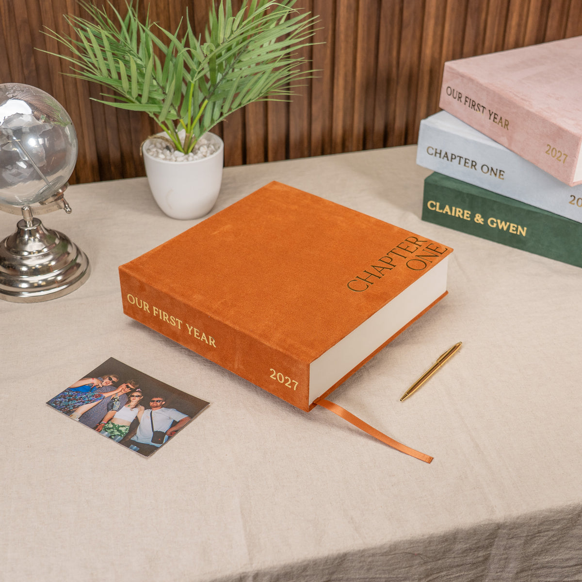An orange hardcover book titled 'Our First Year 2027' sits on a beige tablecloth, styled with a photo print, gold pen, potted plant, and a stack of books in the background.