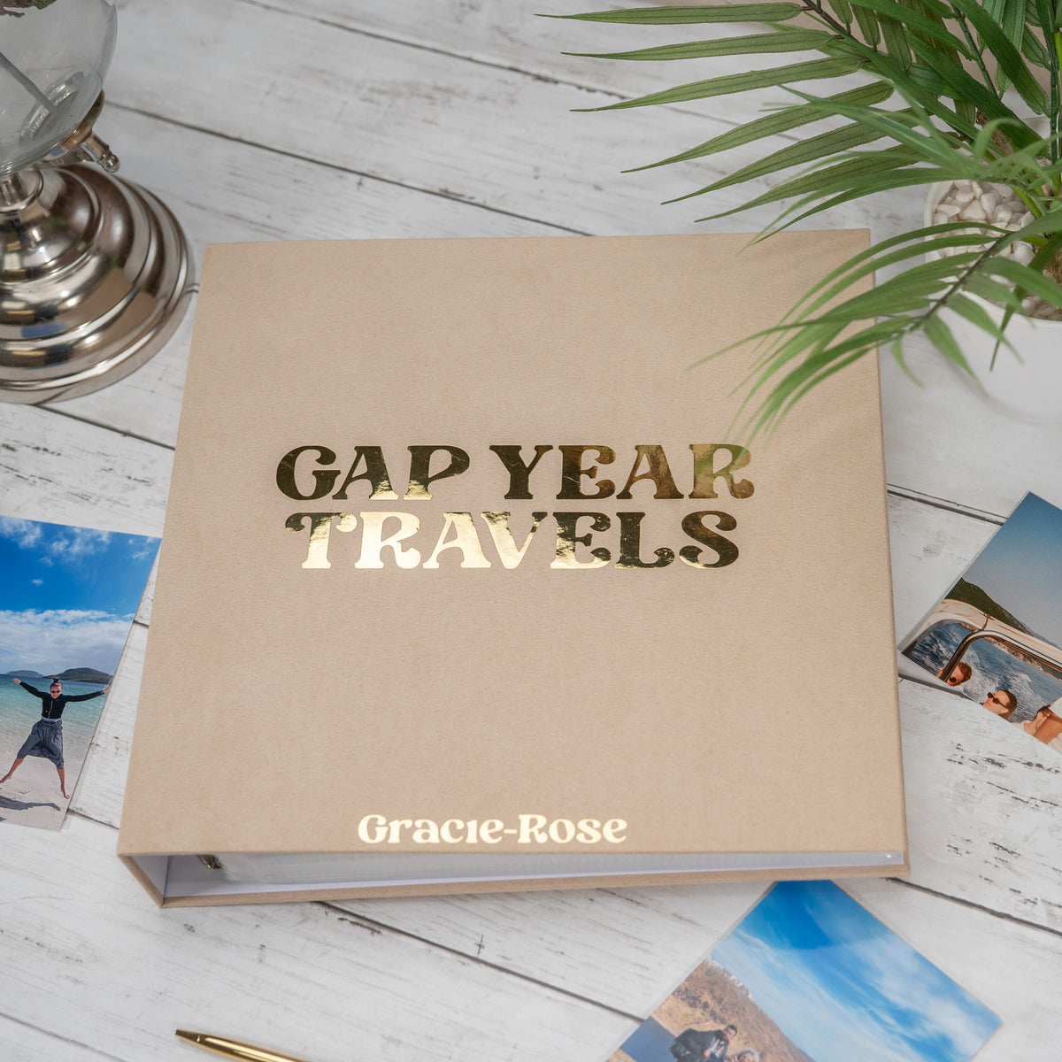 A beige leather-bound travel photo album with 'GAP YEAR TRAVELS' in metallic gold foil on the cover, styled on a white wooden surface with photos, a plant, and a small lamp.