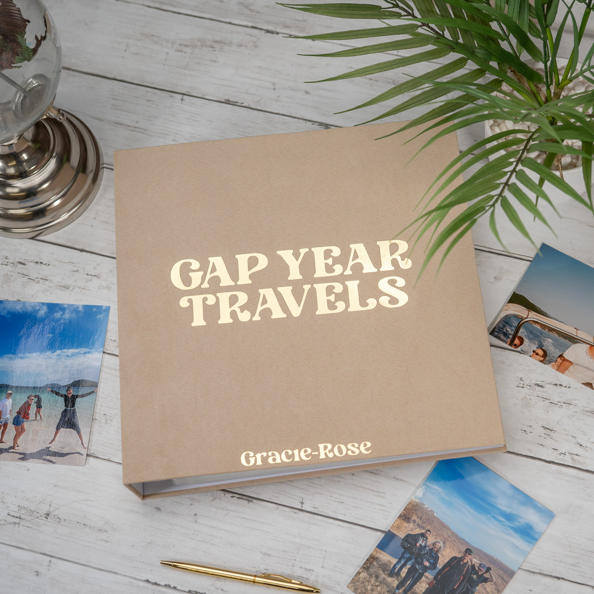 A kraft paper travel photo album titled 'GAP YEAR TRAVELS' by Gracie-Rose, styled on a white wooden tabletop with scattered travel photos, a gold pen, and a green plant.