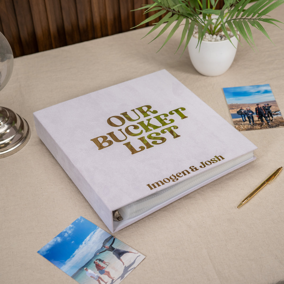 A white hardcover photo album labeled 'OUR BUCKET LIST' with the names 'Imogen & Josh' in gold foil, placed on a beige tablecloth with small printed photos, a pen, and a potted plant around it.