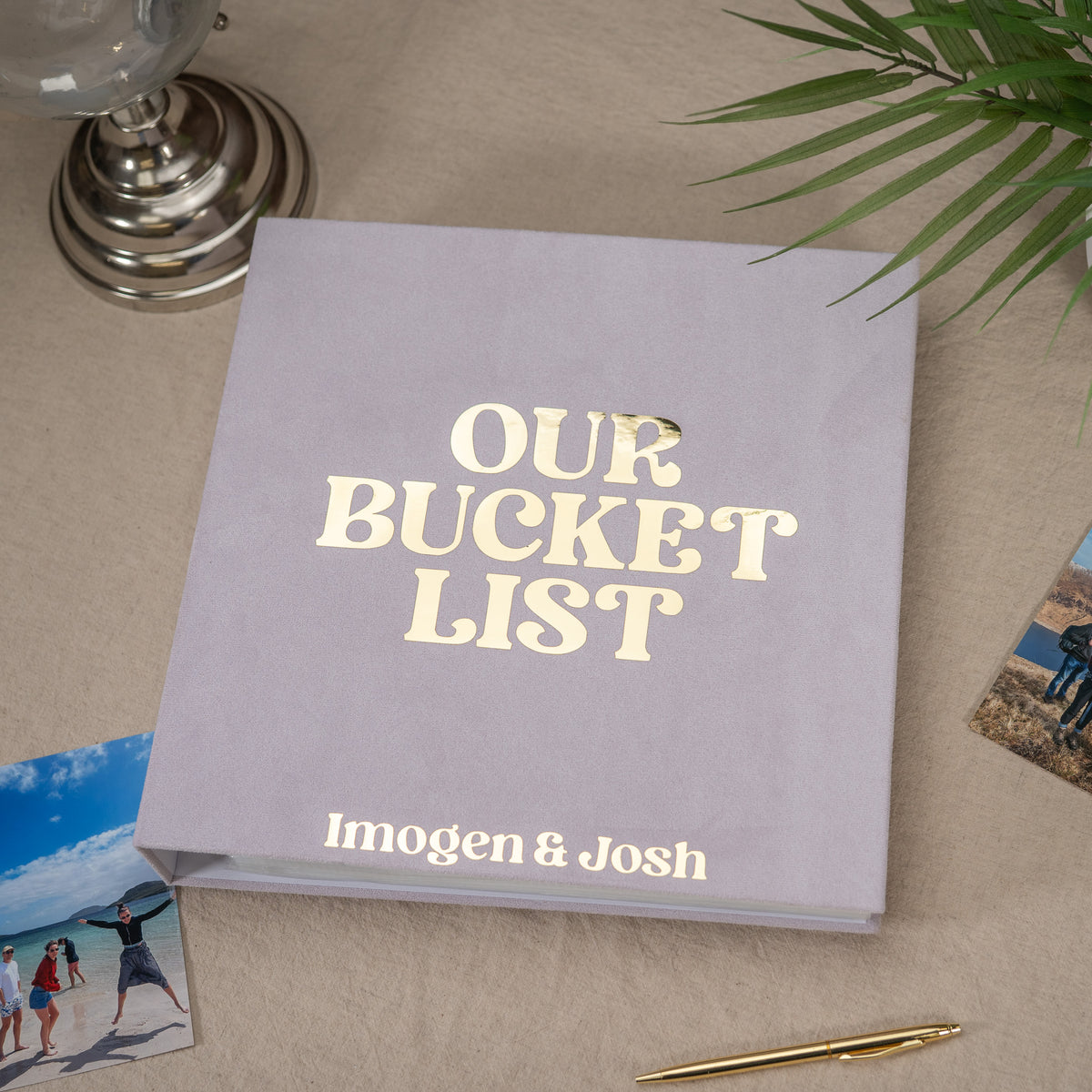 A personalized photo album titled 'OUR BUCKET LIST' with the names 'Imogen & Josh' printed on the front, styled on a beige fabric surface with a glass lamp base, plant leaves, and small photos around it.