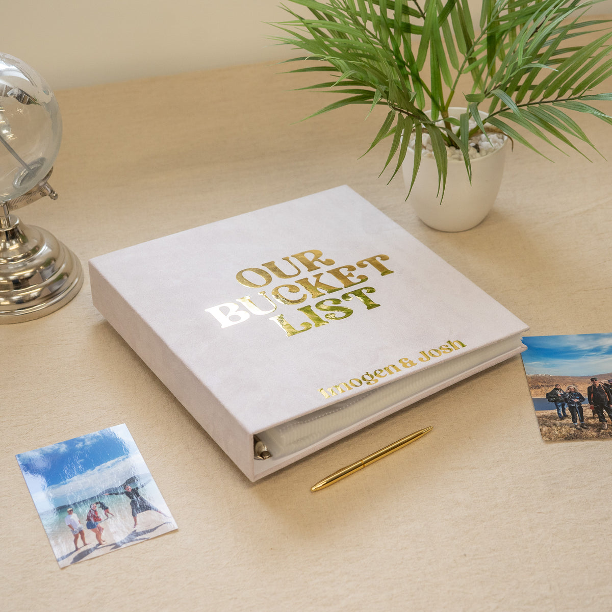 A white hardcover binder with gold foil text reading 'OUR BUCKET LIST' and the names 'Maggie & Josh' sits on a light wood desk, styled with a gold pen, small printed photos, and a potted plant.