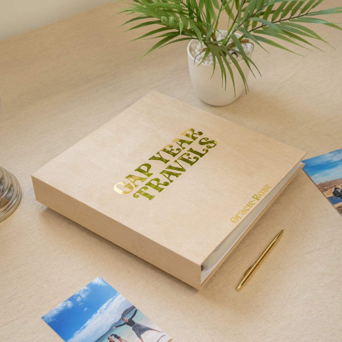 A beige hardcover travel photo album titled 'GAP YEAR TRAVELS' with gold foil lettering, placed on a light wood desk next to a small potted plant, a gold pen, and printed photos.