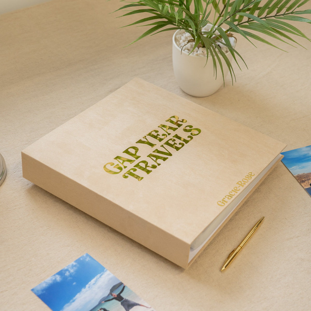 A beige hardcover photo album titled 'GAP YEAR TRAVELS' in gold foil, placed on a light wood desk with a small potted plant, a gold pen, and travel photos nearby.