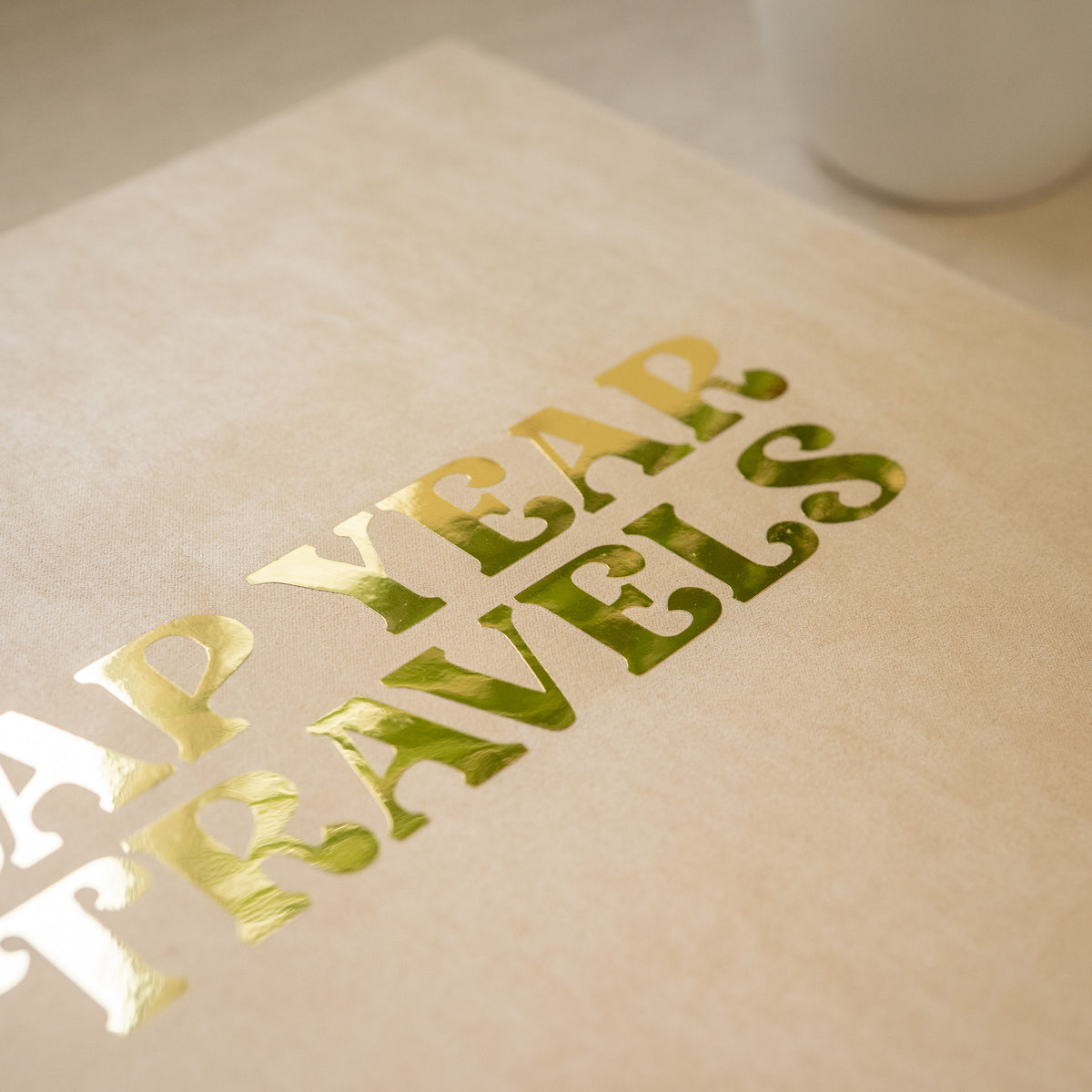 A close-up of a beige textured paper item featuring metallic gold foil cutout text that reads 'HAPPY YEAR TRAVELS' diagonally.