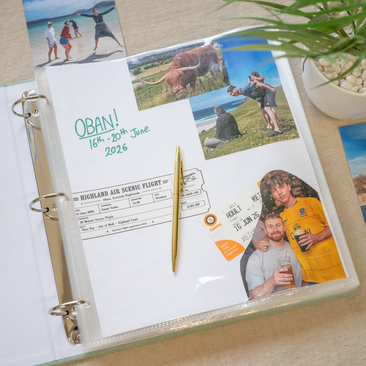 A clear plastic ring-bound photo album open on a light beige surface, showing a decorated travel-themed page with printed photos, handwritten text, ticket-style graphics, and a gold pen. A plant leaf and part of another card are visible around it.