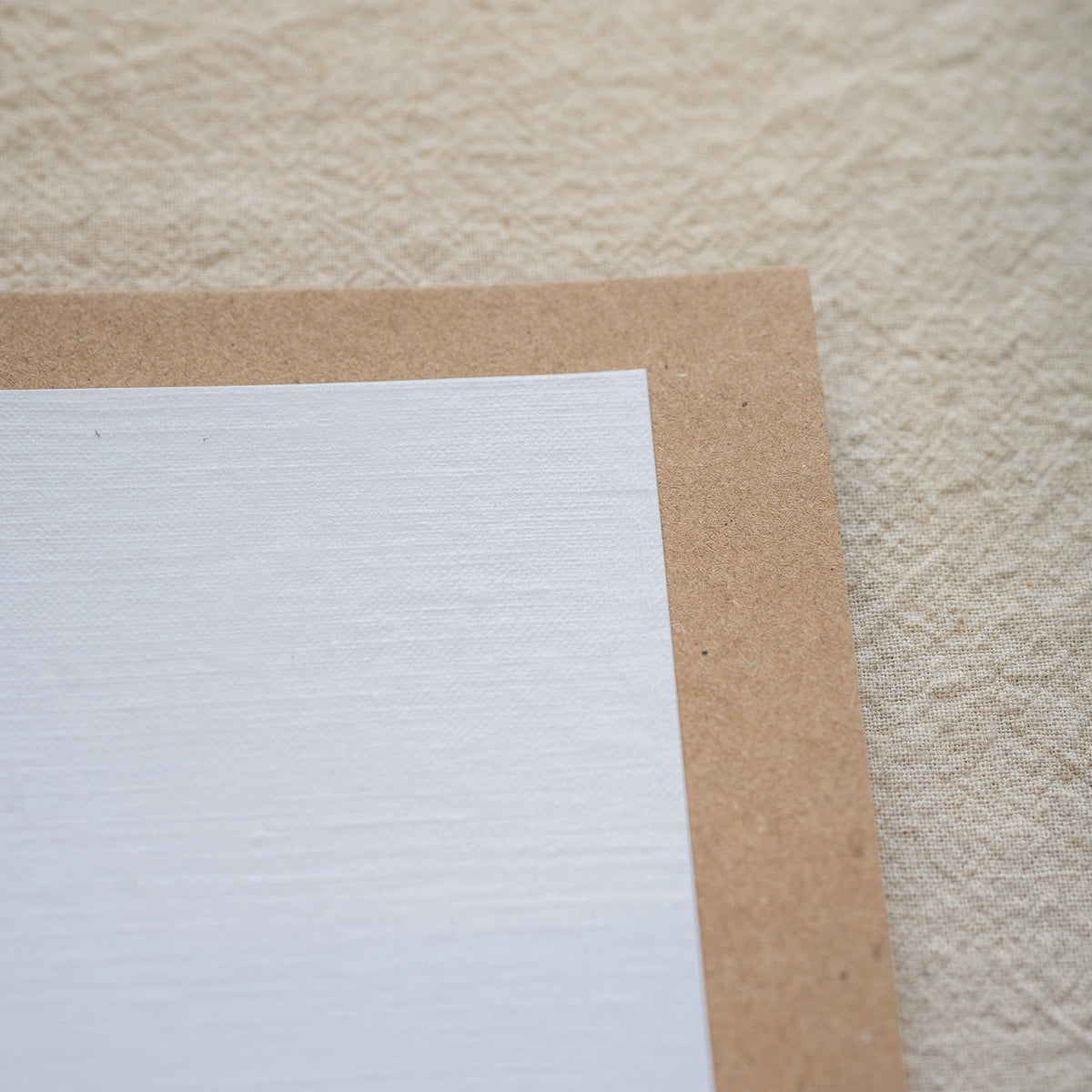 A close-up of a white textured paper sheet with a brown kraft paper border, placed on a beige textured fabric background.