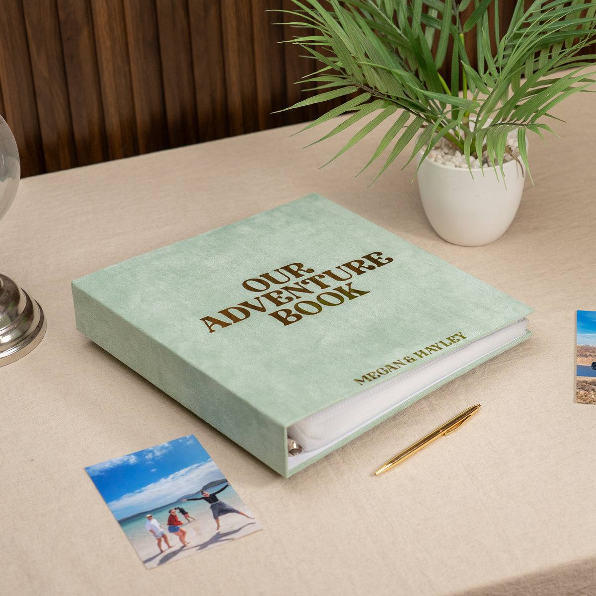 A personalized hardcover photo album titled 'OUR ADVENTURE BOOK' with the name 'MEGAN HARTLEY' printed on the front, styled on a beige table next to a potted plant, a pen, and printed photos.