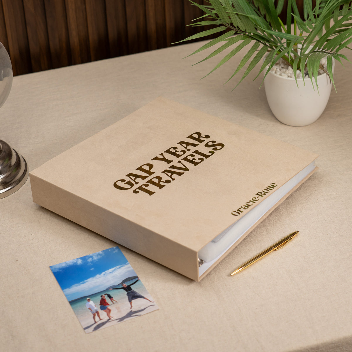 A beige Gap Year Travels binder with custom text, styled on a linen tablecloth next to a photo print, a gold pen, and a potted plant.