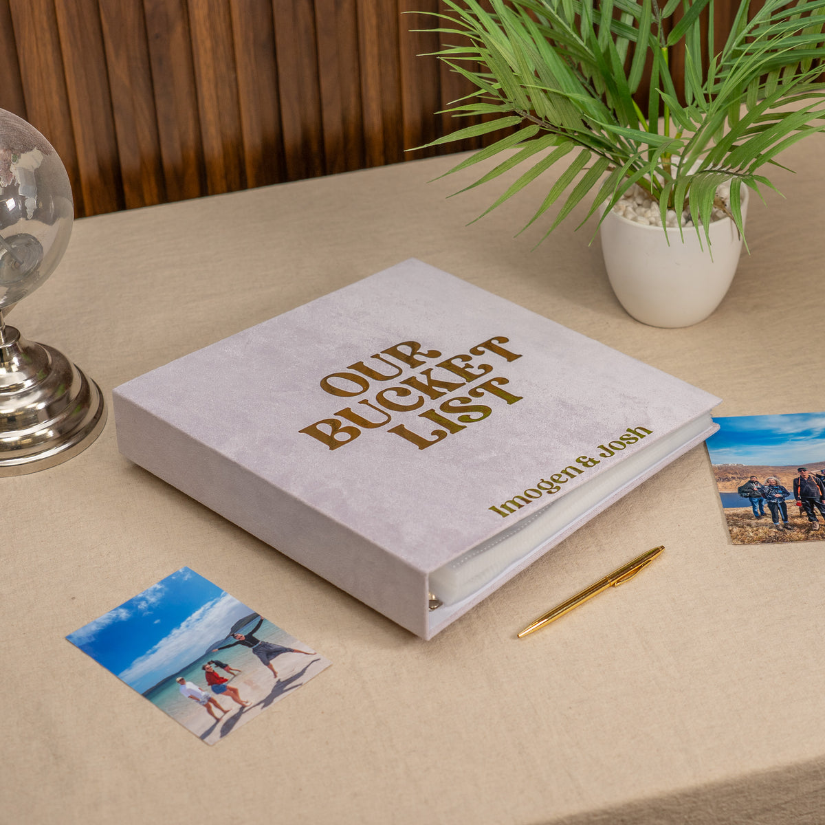 A light gray binder labeled 'OUR BUCKET LIST' sits on a beige tablecloth with printed photos, a gold pen, and a potted plant in the background.