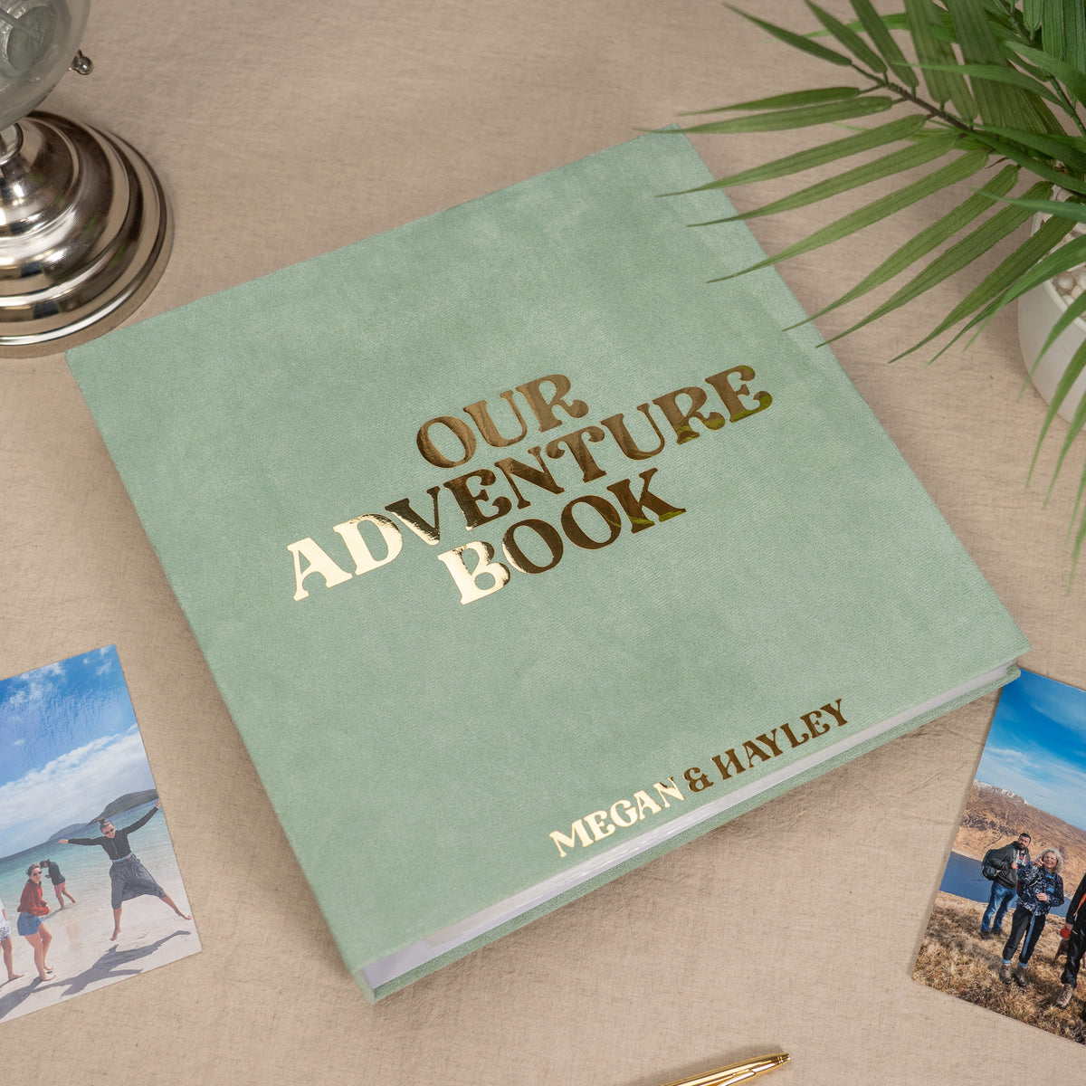 A personalized mint green hardcover photo book titled 'OUR ADVENTURE BOOK' with gold foil lettering, displayed on a beige fabric surface with small printed photos and a plant leaf around it.