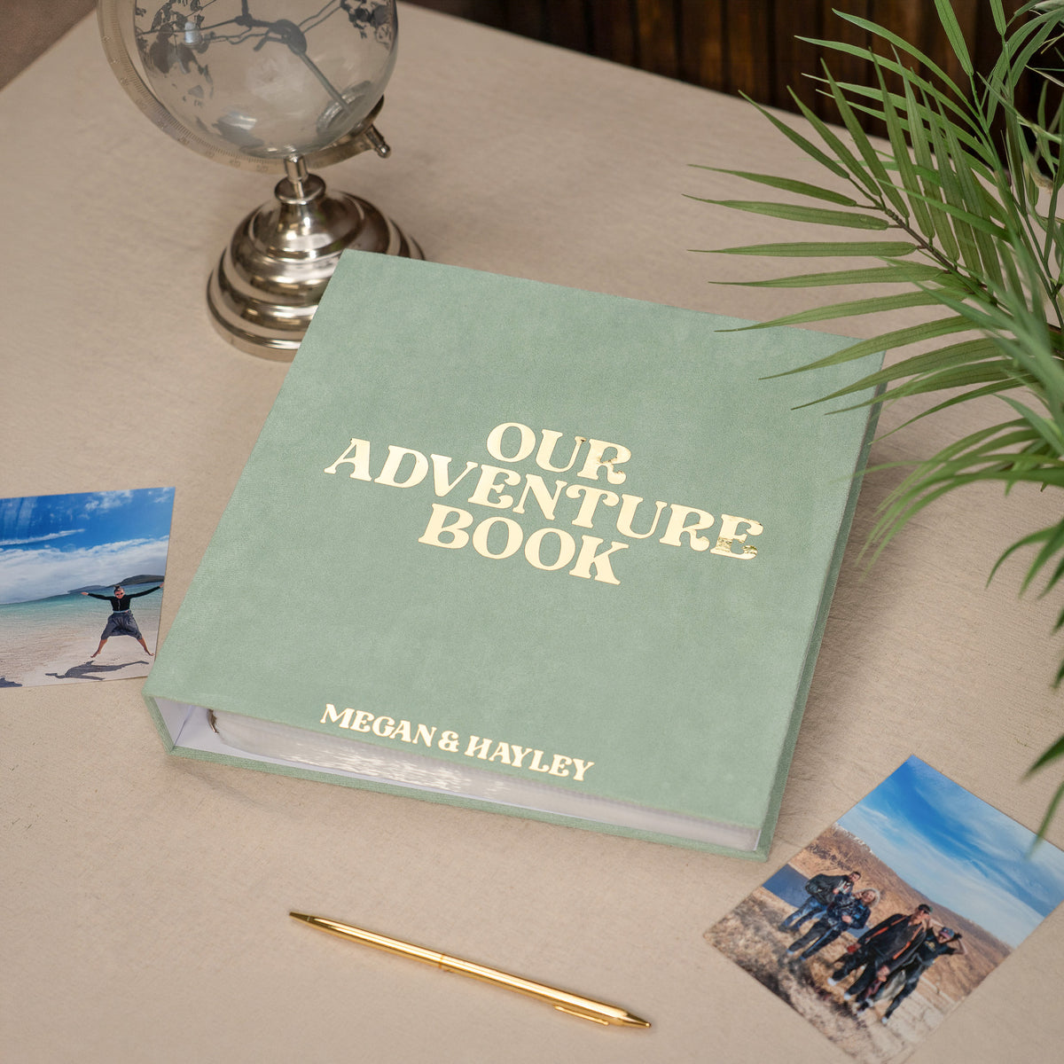 A personalized hardcover photo album titled 'OUR ADVENTURE BOOK' with the names 'MEGAN & HAYLEY' printed on the front, styled on a beige tabletop with a gold pen, beach photos, a globe, and a plant.