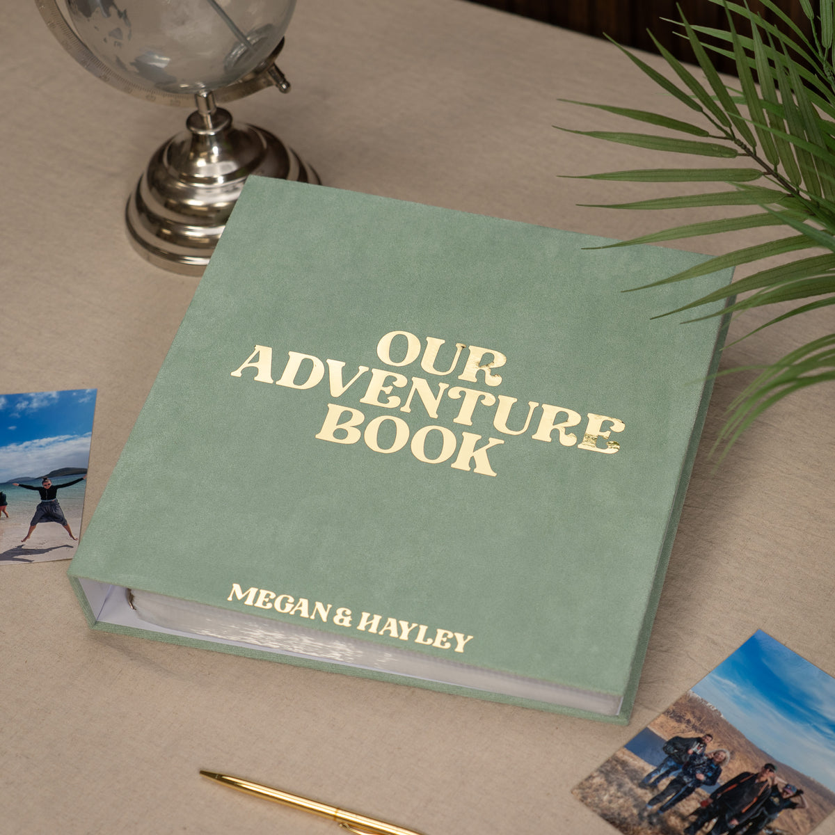 A personalized hardcover 'Our Adventure Book' in sage green with gold foil lettering, styled on a beige tablecloth with a globe, pen, and beach photos.