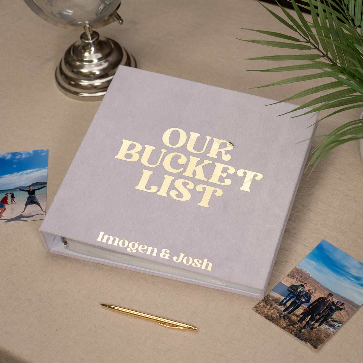A personalized hardcover photo album titled 'OUR BUCKET LIST' with the names 'Imogen & Josh' printed on the front, styled on a beige tabletop with a gold pen, small photos, and a plant.