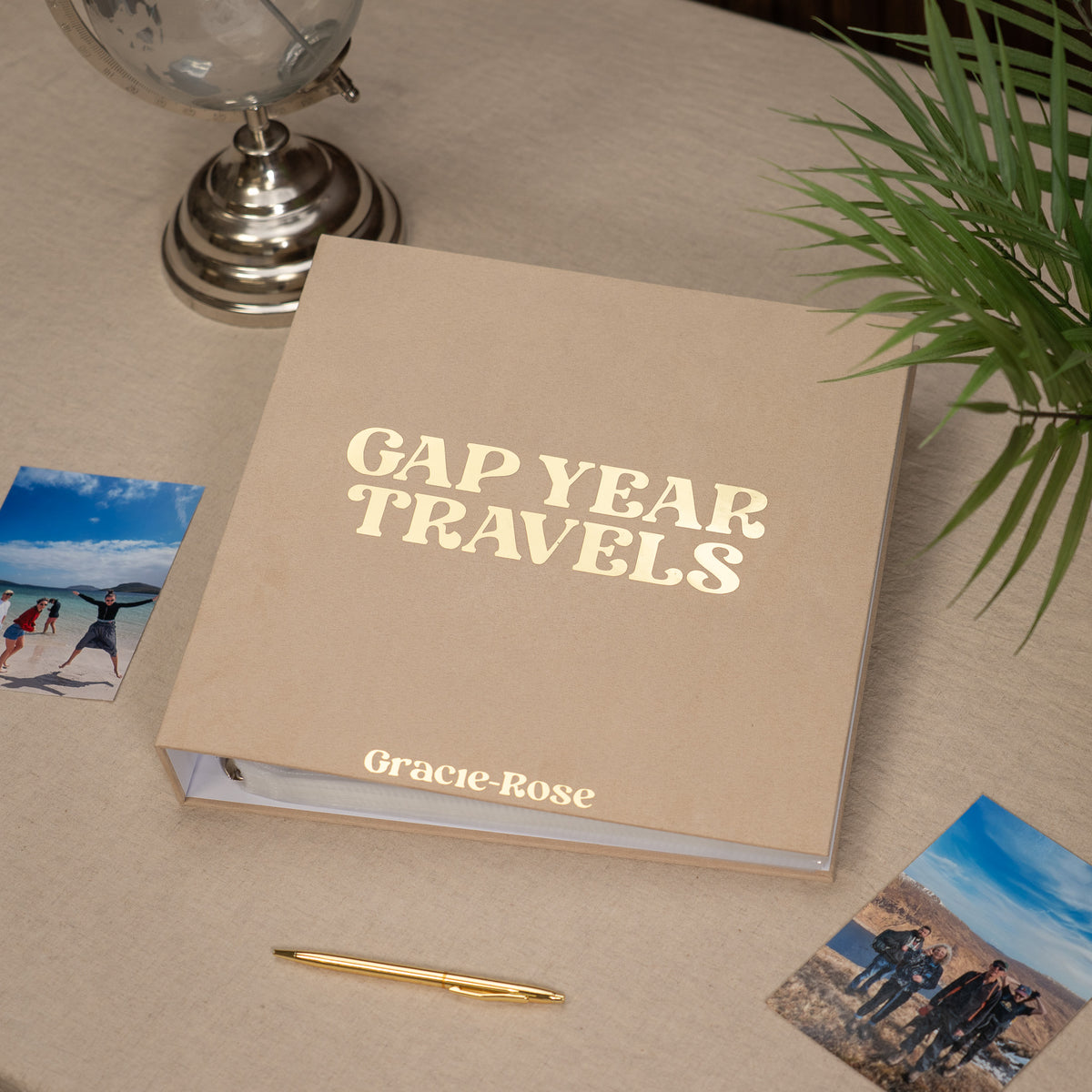 A beige hardcover travel photo album titled 'GAP YEAR TRAVELS' with the brand name 'Gracie-Rose' printed on the front, styled on a beige tabletop with a gold pen, small travel photos, a plant, and a globe in the background.