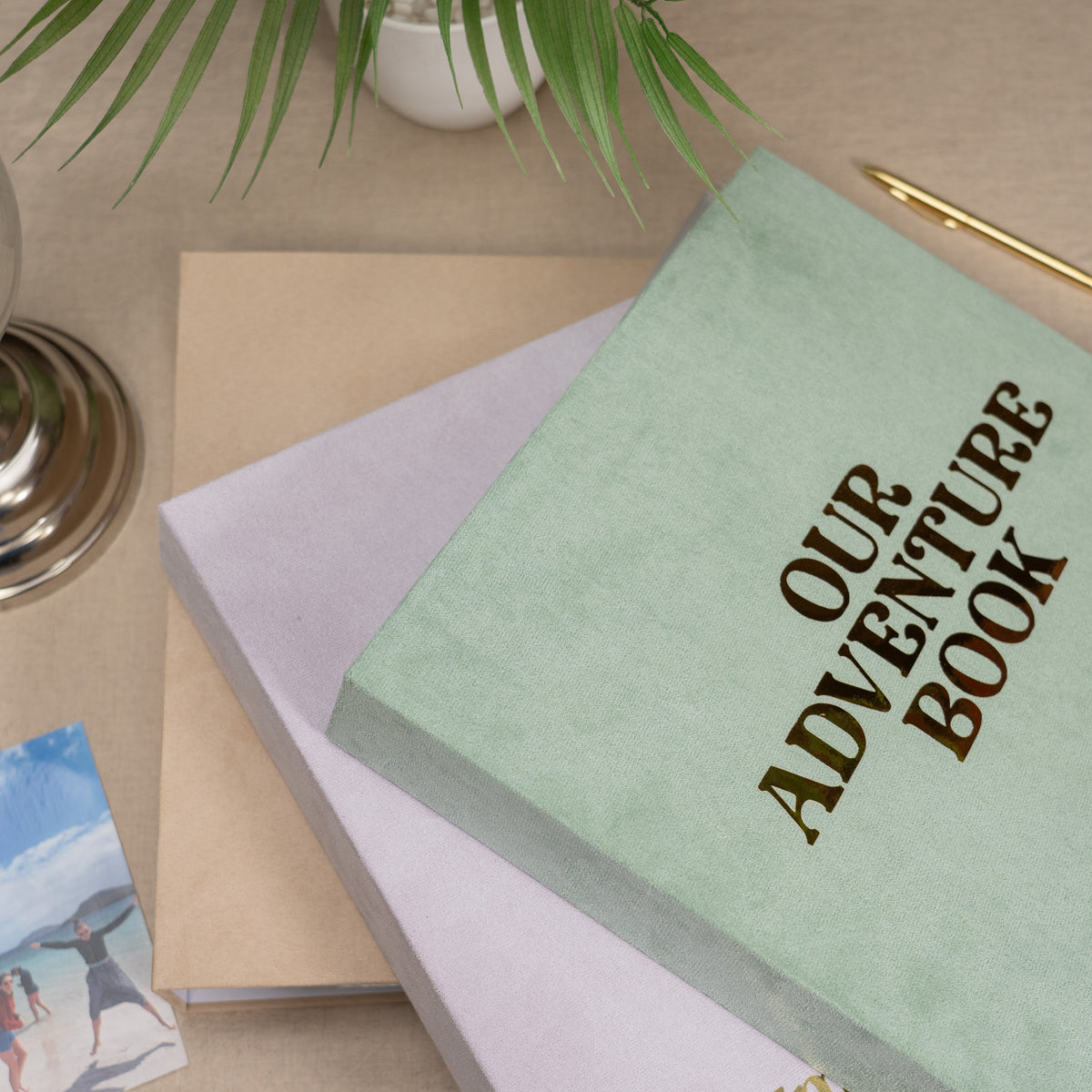 A styled flat-lay of a hardcover journal or notebook with a sage green cover reading 'OUR ADVENTURE BOOK' in dark lettering, layered over a light lavender book and a kraft paper book on a beige tabletop with decorative plant leaves and a gold pen.