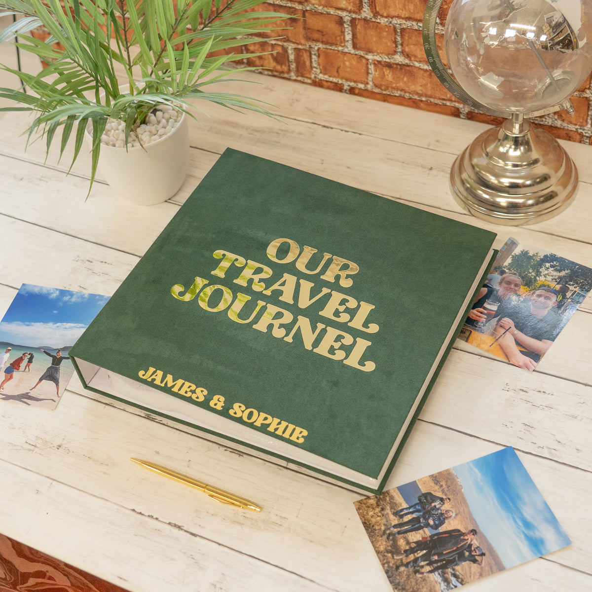 A personalized dark green hardcover travel journal with gold foil lettering reading 'OUR TRAVEL JOURNAL' and 'JAMES & SOPHIE', styled on a white wooden table with photos, a plant, and a brass globe.