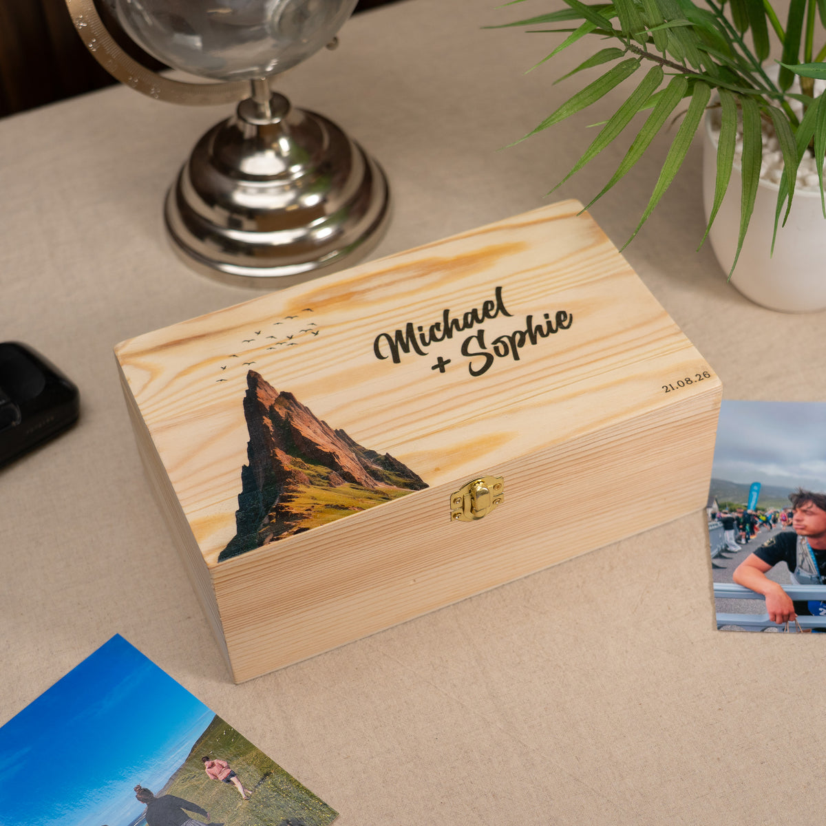 A personalized wooden keepsake box with a mountain illustration and the names 'Michael + Sophie' on the lid, placed on a beige tabletop with photos and decor in the background.