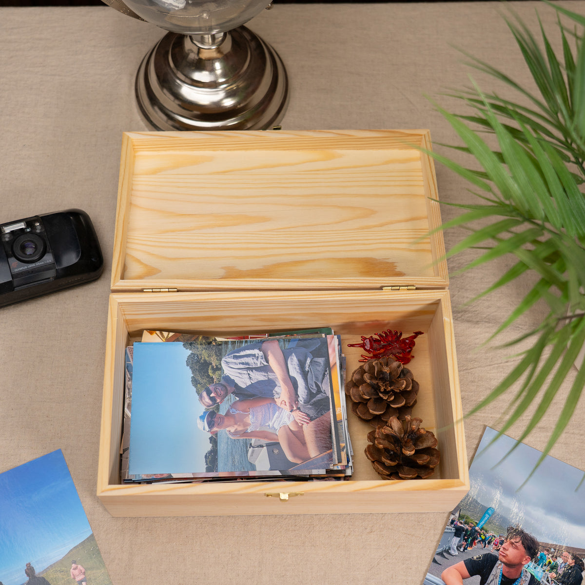 A light natural wood photo box with its lid open, shown on a beige tabletop. Inside the box are printed photographs, pinecones, and a small red decorative item.