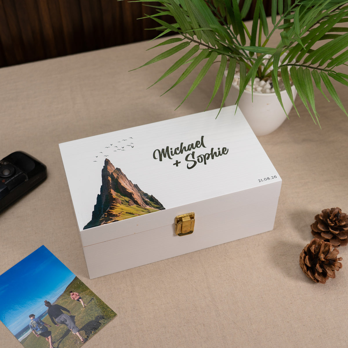 A personalized white wooden keepsake box with a gold clasp, featuring a printed mountain illustration and the names 'Michael + Sophie' on the lid. The box is styled on a beige fabric surface with a potted plant, pinecones, a photo print, and a black remote nearby.