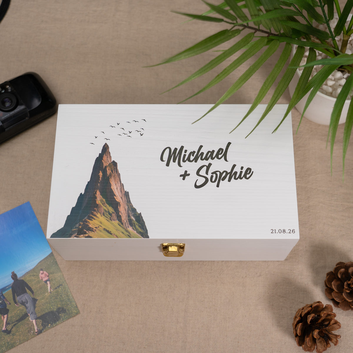 A personalized white wooden keepsake box with a gold clasp, featuring a printed mountain illustration and the names 'Michael + Sophie' on the lid. The box is styled on a beige fabric surface with a plant, pinecones, and a photo card nearby.