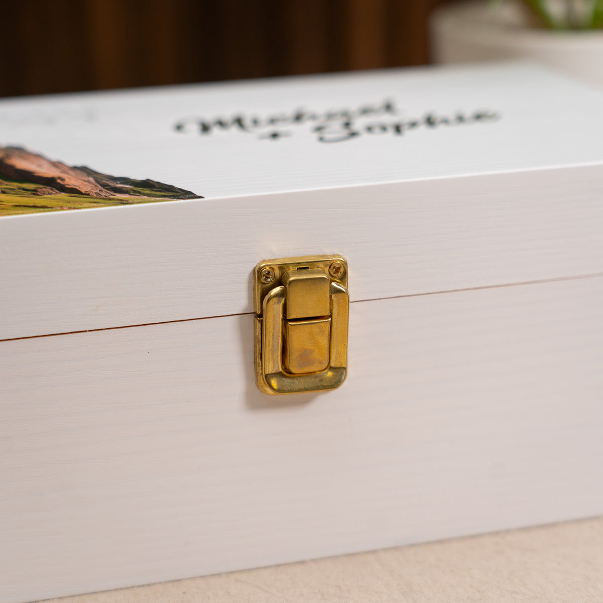A close-up of a white wooden keepsake box with a gold metal latch clasp, featuring custom printed names 'Michael & Sophie' and a landscape illustration on the lid.