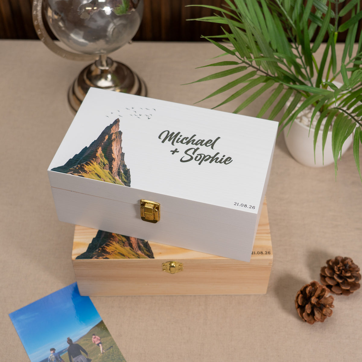 A personalized wooden keepsake box with a white lid featuring a mountain illustration and the names 'Michael + Sophie' in script, displayed on top of a natural wood box. The scene is styled with a globe, a potted palm, and pinecones on a beige surface.
