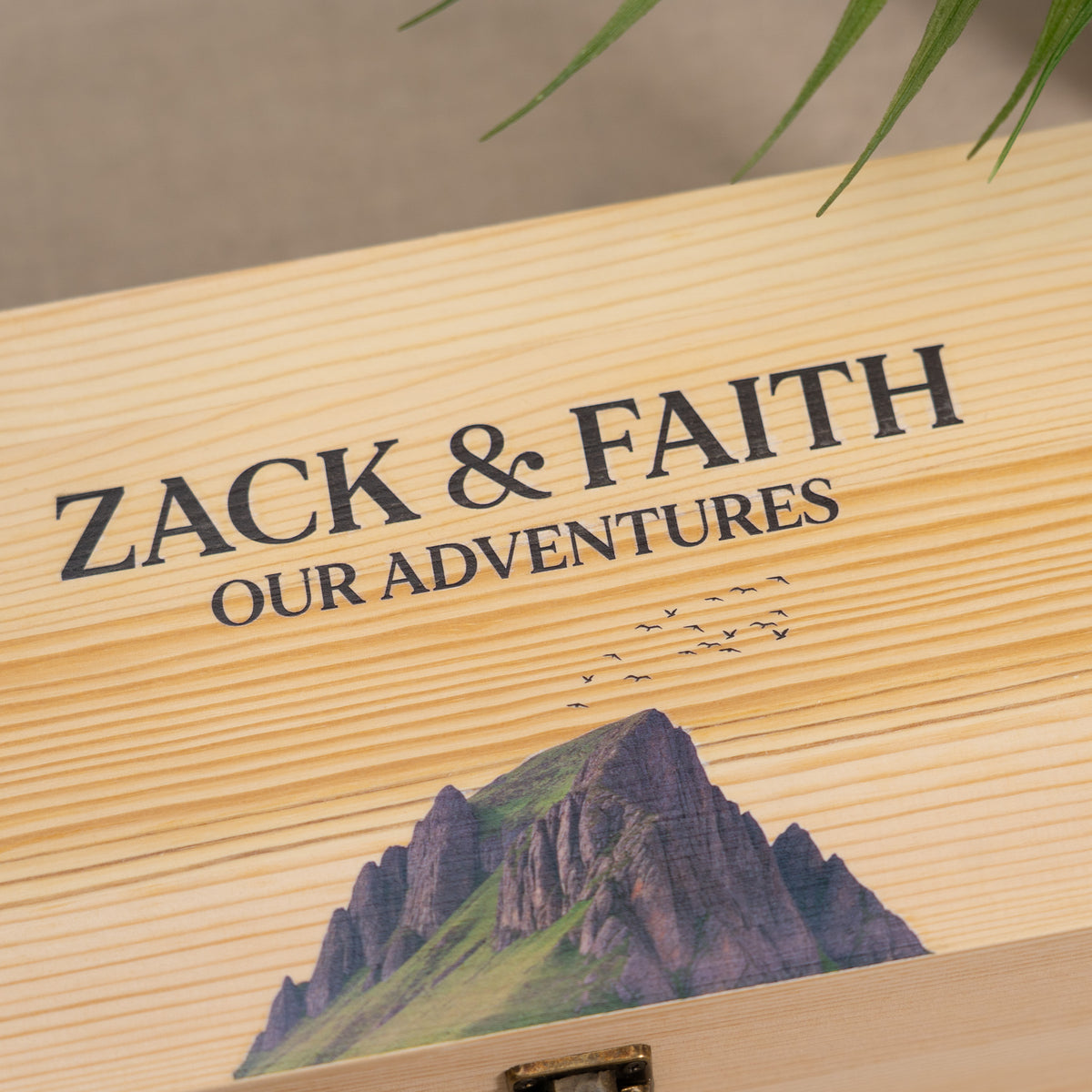Close-up of a light natural wood box lid engraved with the words 'ZACK & FAITH OUR ADVENTURES' above a mountain illustration, with a small metal latch visible at the bottom.