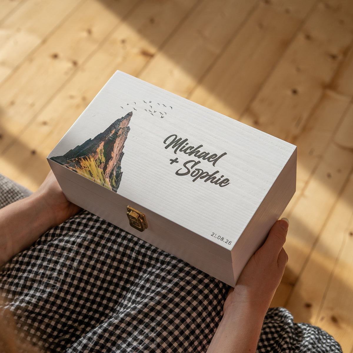 A person holds a personalized white wooden keepsake box with a gold clasp. The lid features a printed mountain illustration and the names 'Michael + Sophie' in elegant script, along with the date '21.
