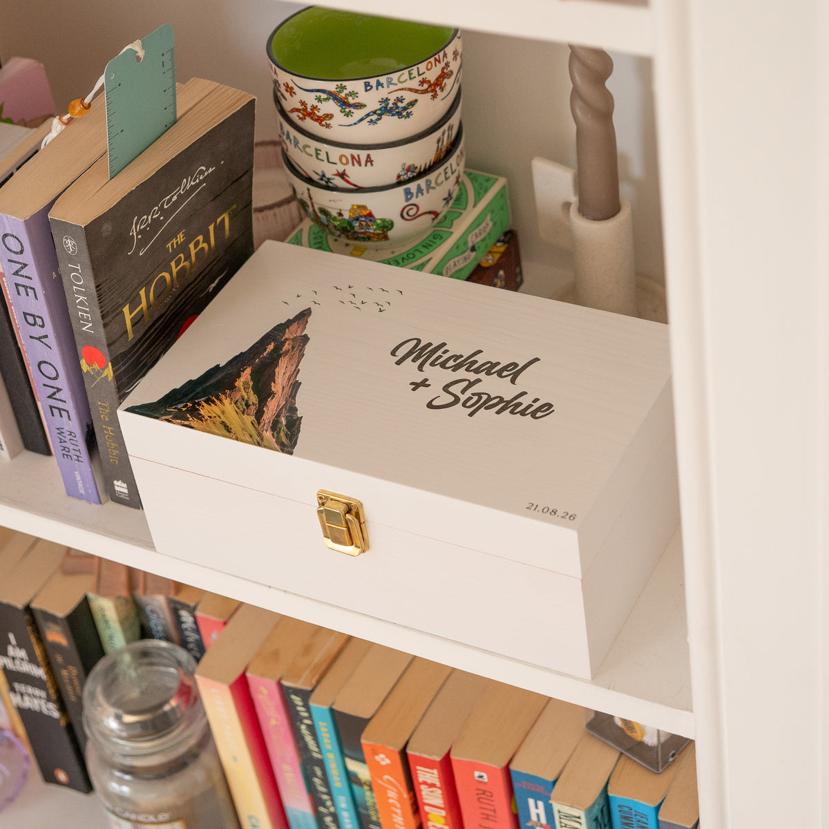 A white personalized keepsake box with a gold clasp sits on a white bookshelf. The lid is printed with the names 'Michael + Sophie' in black script, alongside a scenic mountain illustration.