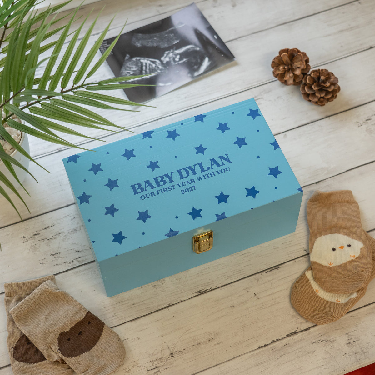 A personalized baby keepsake box in light blue with dark blue stars and the text 'BABY DYLAN OUR FIRST YEAR WITH YOU 2027', styled on a white wooden surface with baby socks, a plant leaf, pinecones, and an ultrasound photo.
