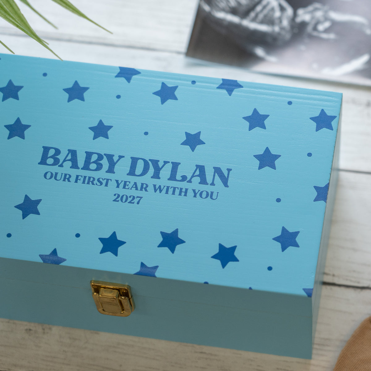 A personalized baby keepsake box in light blue with dark blue star patterns and the text 'BABY DYLAN OUR FIRST YEAR WITH YOU 2027' on the lid, shown on a white wood surface with a blurred photo in the background.