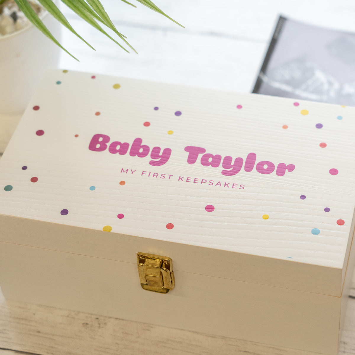 A personalized baby keepsake box with a white wood-grain finish, decorated with colorful confetti dots and the text 'Baby Taylor' and 'MY FIRST KEEPSAKES' on the lid. The box features a gold clasp and is shown in a styled setting with a plant leaf in the foreground.