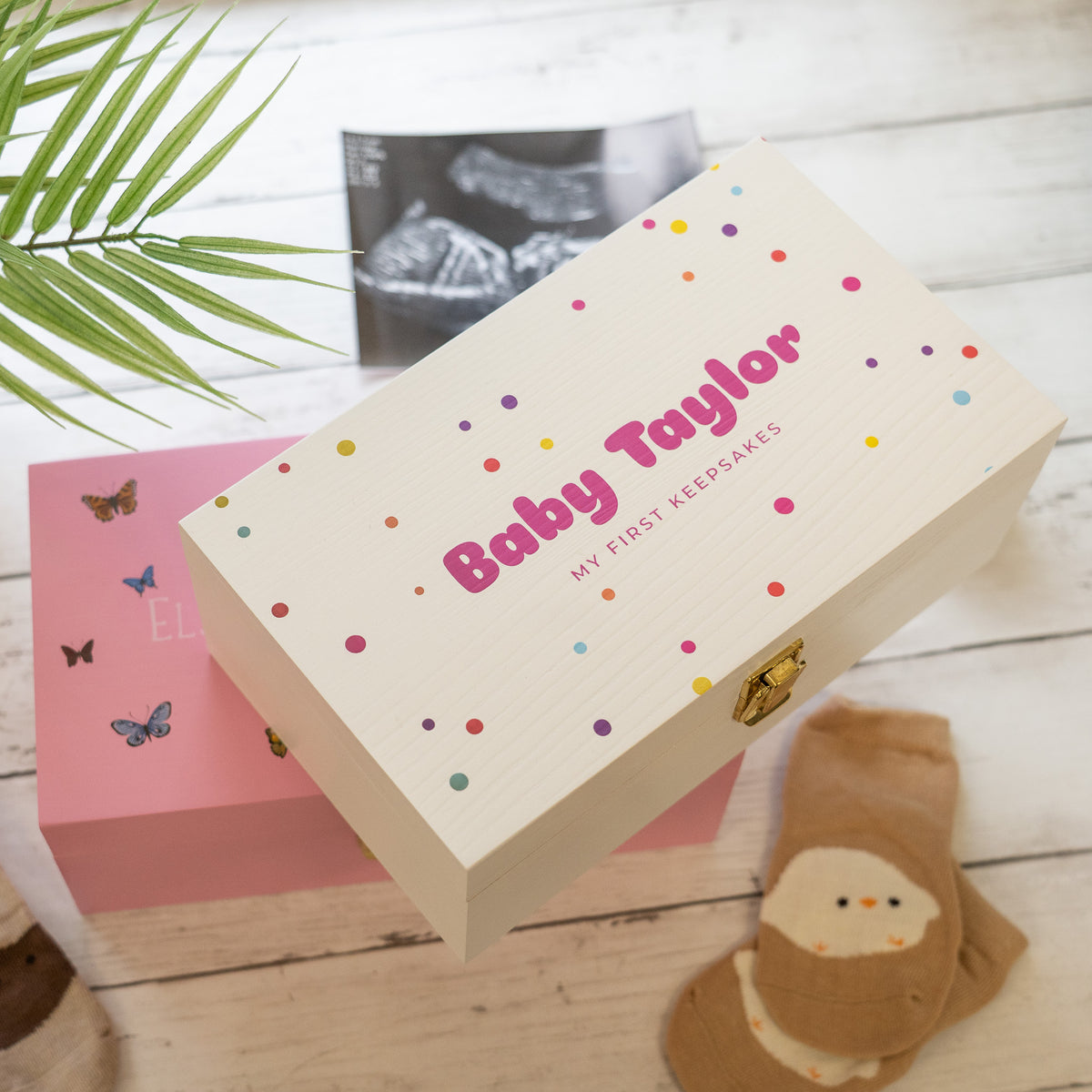 A personalized cream-colored wooden baby keepsake box with colorful polka dots and pink text reading 'Baby Taylor MY FIRST KEEPSAKES', styled on a white wood surface with a pink box underneath, baby socks, and a plant leaf.