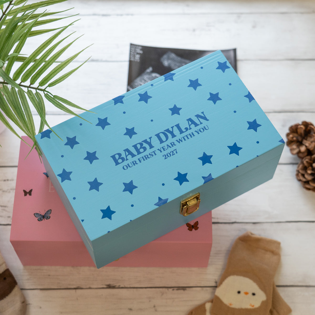 A personalized baby keepsake box in light blue with dark blue stars and the text 'BABY DYLAN OUR FIRST YEAR WITH YOU 2027', displayed on top of a pink box on a white wood background with decorative props.