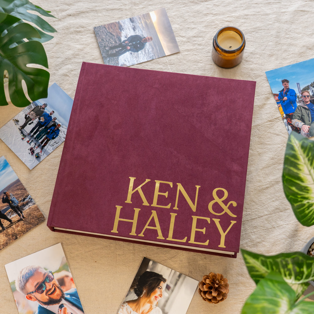 A personalized hardcover photo album with a deep burgundy cover and gold foil lettering reading 'KEN & HALEY', styled in a flat lay on a cream fabric background with scattered photos, green leaves, a candle, and a pine cone.