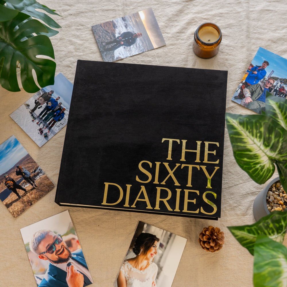 A black velvet photo album titled 'THE SIXTY DIARIES' displayed on a table with an arrangement of photos, pine cones, and a candle.