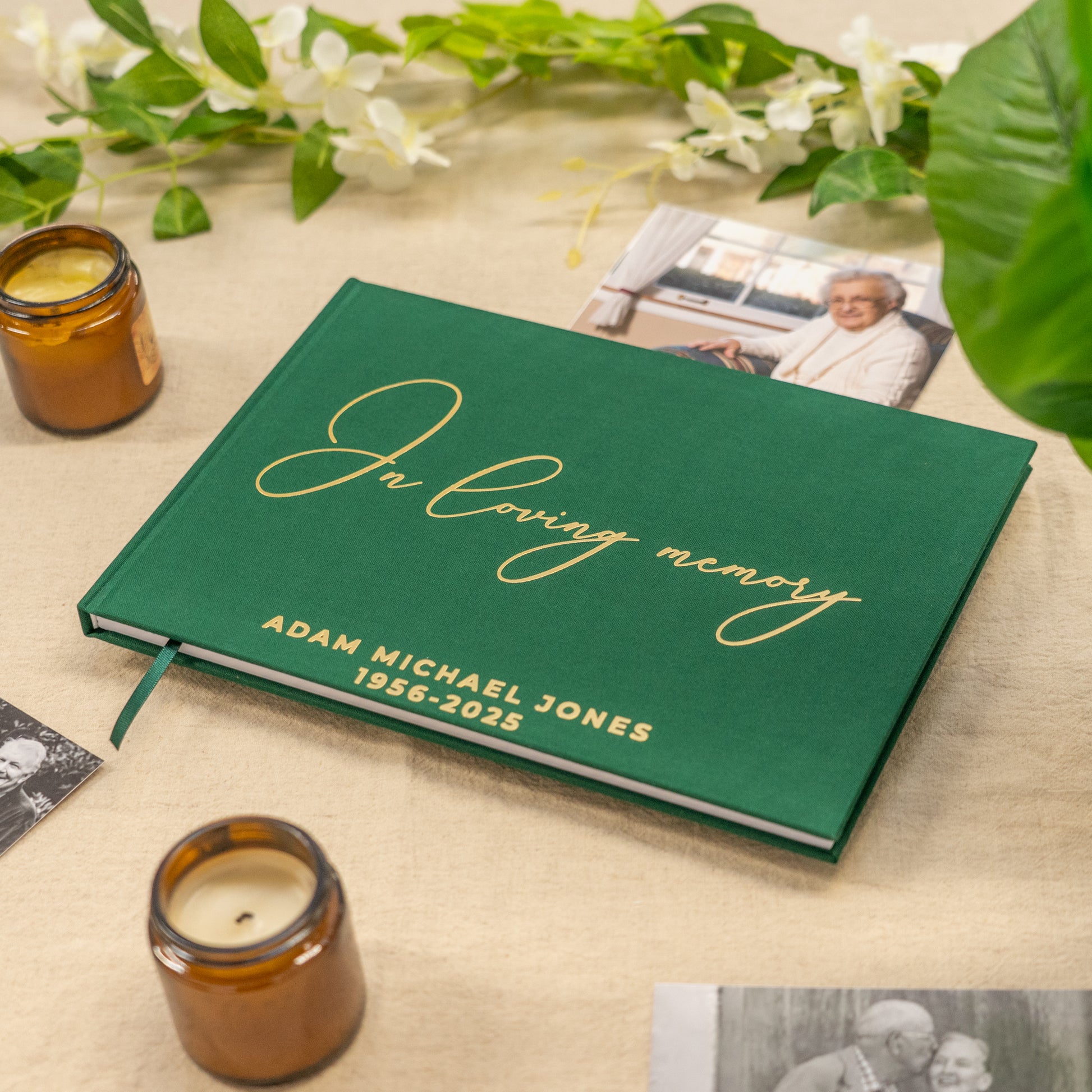 Green book titled 'In Loving Memory' with gold text on a beige surface with candles and photos. condolence book