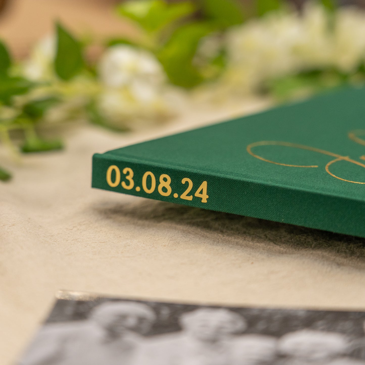 Green book with gold text '03.08.24' on a surface with blurred greenery and a black and white photo in the background.