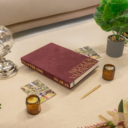 Maroon book titled 'Chapter One' with personalized text on a beige surface, surrounded by candles and a plant.