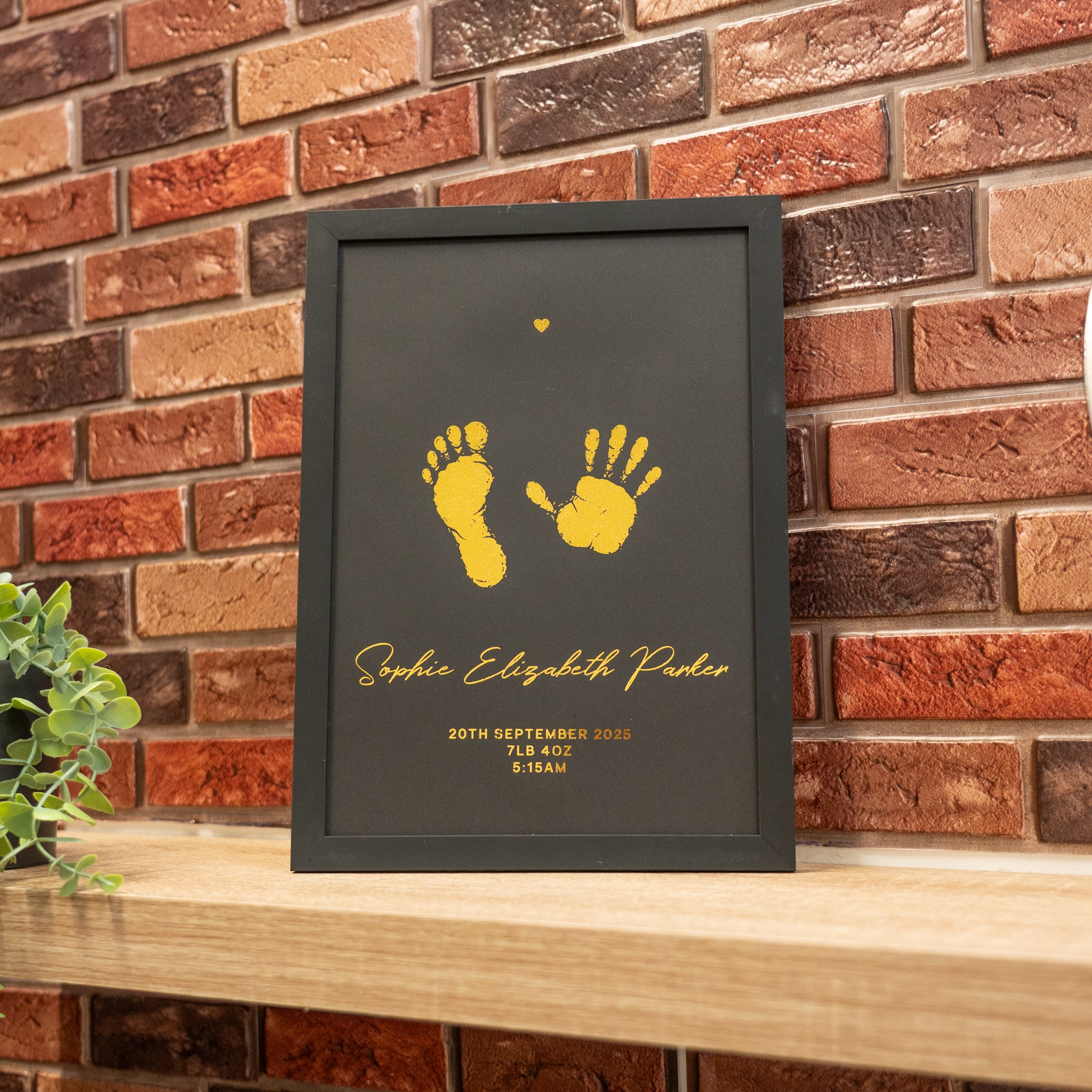 Framed print with yellow footprints and handprints on a dark background, leaning against a brick wall.