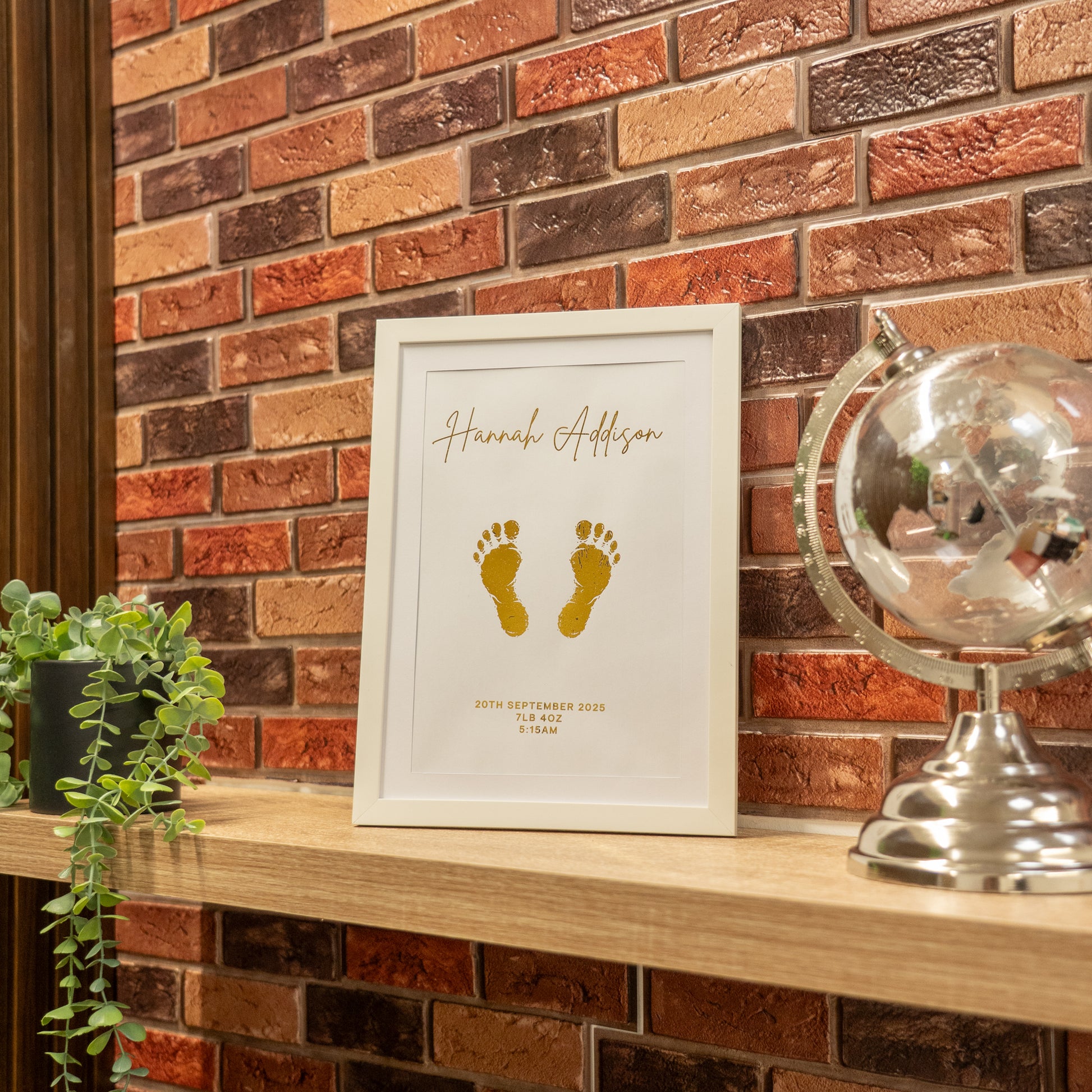Framed baby footprint artwork on a wooden shelf against a brick wall.
